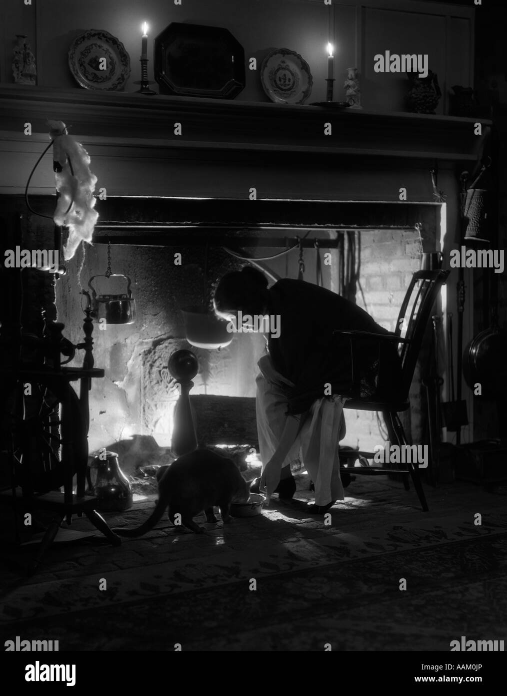 1920ER JAHREN FRAU KAMINFEUER SITZEN IM STUHL, BEUGTE SICH ÜBER INDEM SCHALE FUTTER KATZE AM BODEN NUR LICHT AUS FEUER & KERZEN Stockfoto