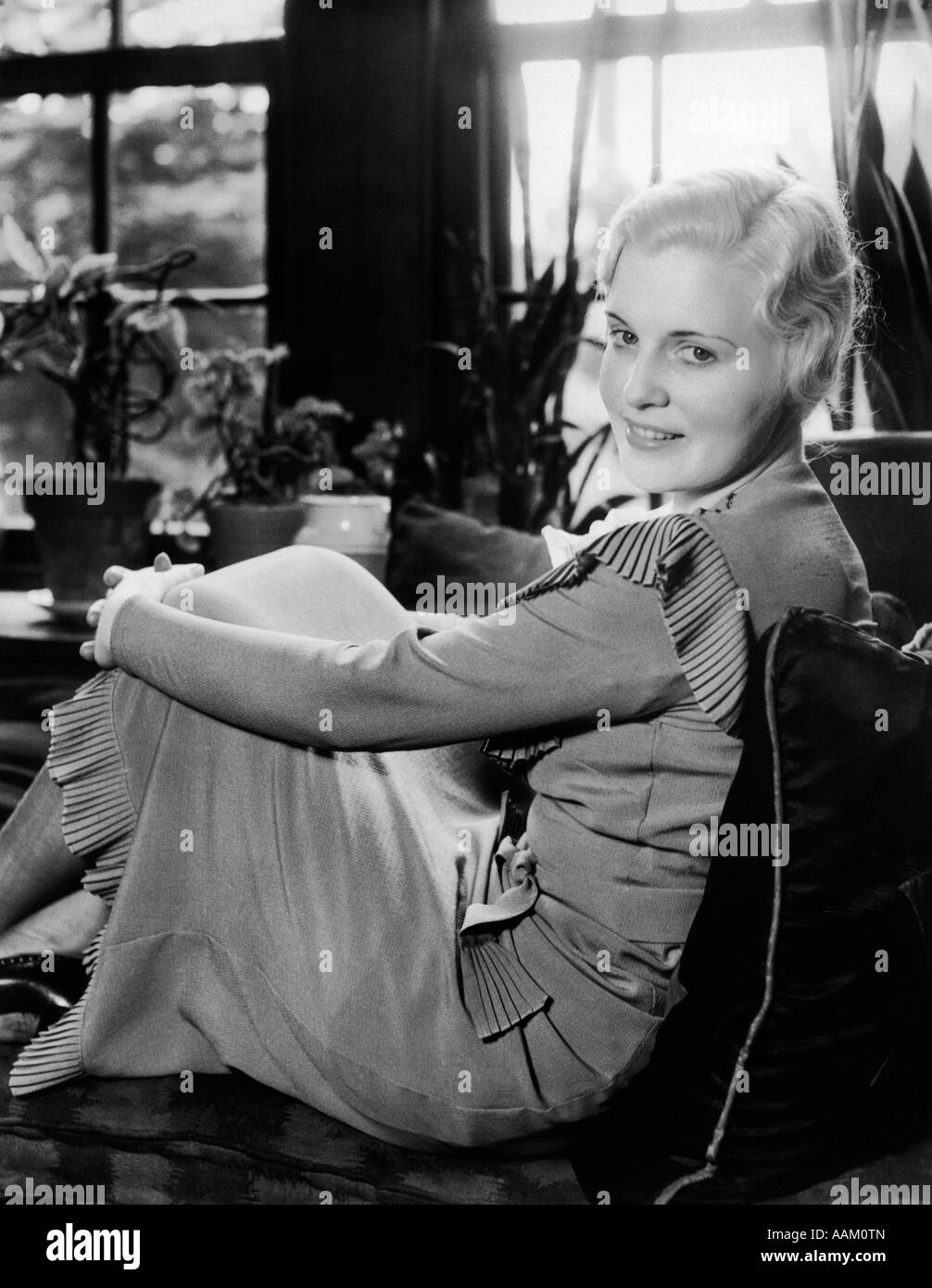 1920S 1930S BLONDE FRAU LÄCHELND MIT SITZEN AUF DEN KNIEN IN FENSTERPLATZ MIT BLICK IN DIE KAMERA ARME Stockfoto