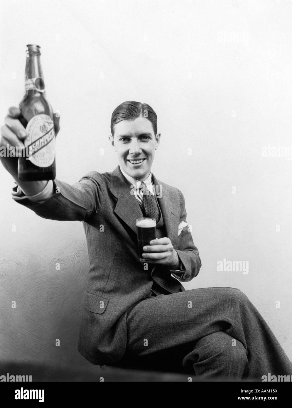1930ER JAHRE MANN HÄLT GROßE VERZERRT BIERFLASCHE IN EINER HAND UND EIN NORMALER GRÖßE GLAS BIER IN ANDEREN LÄCHELNDEN BLICK IN DIE KAMERA Stockfoto