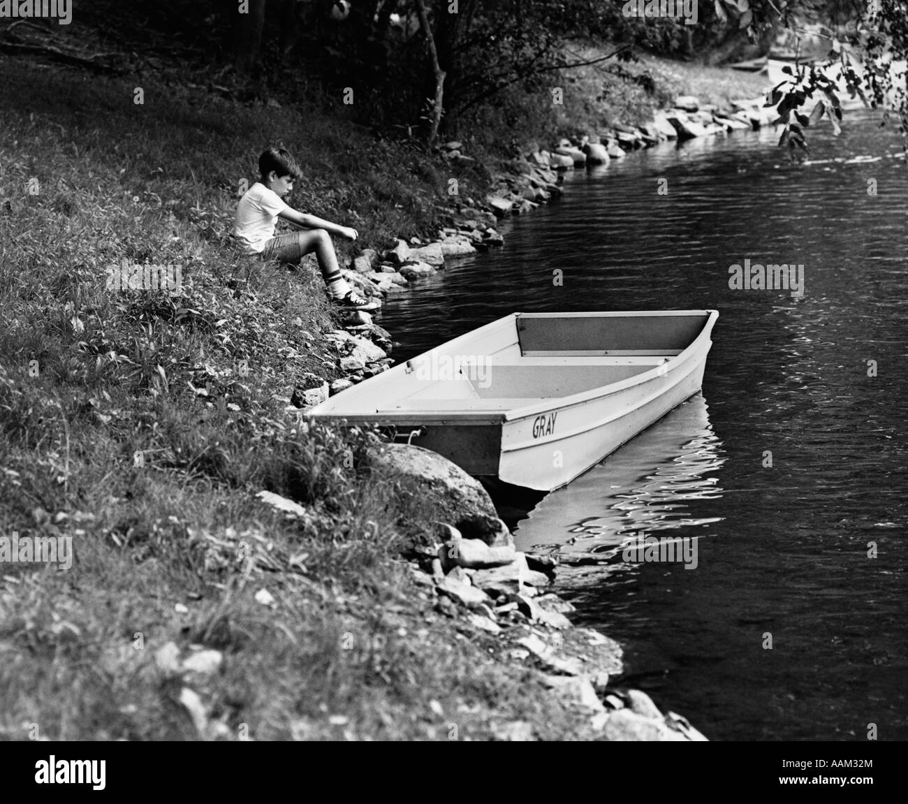 SEITENANSICHT DER 1960ER JAHRE BOY SITTING ON BANK SEE NEBEN RUDERBOOT IM WASSER Stockfoto