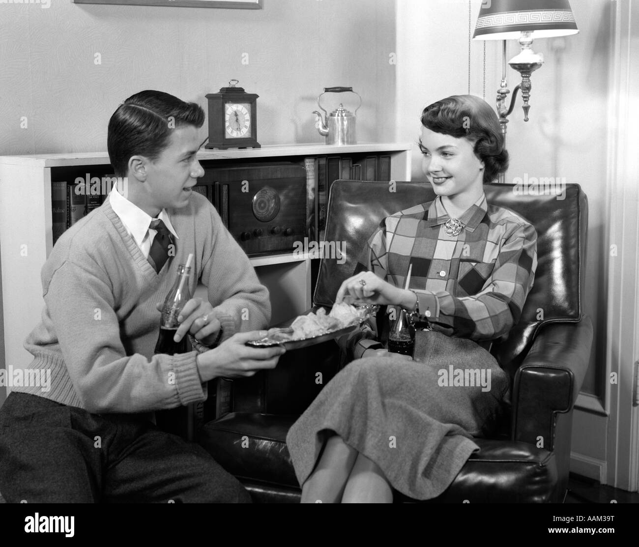 1950ER JAHRE JUNGE MÄDCHEN JUNGE TEEN PAAR AUS DEM SITZEN IM STUHL TRINKEN KOKSEN ESSEN KARTOFFELCHIPS Stockfoto