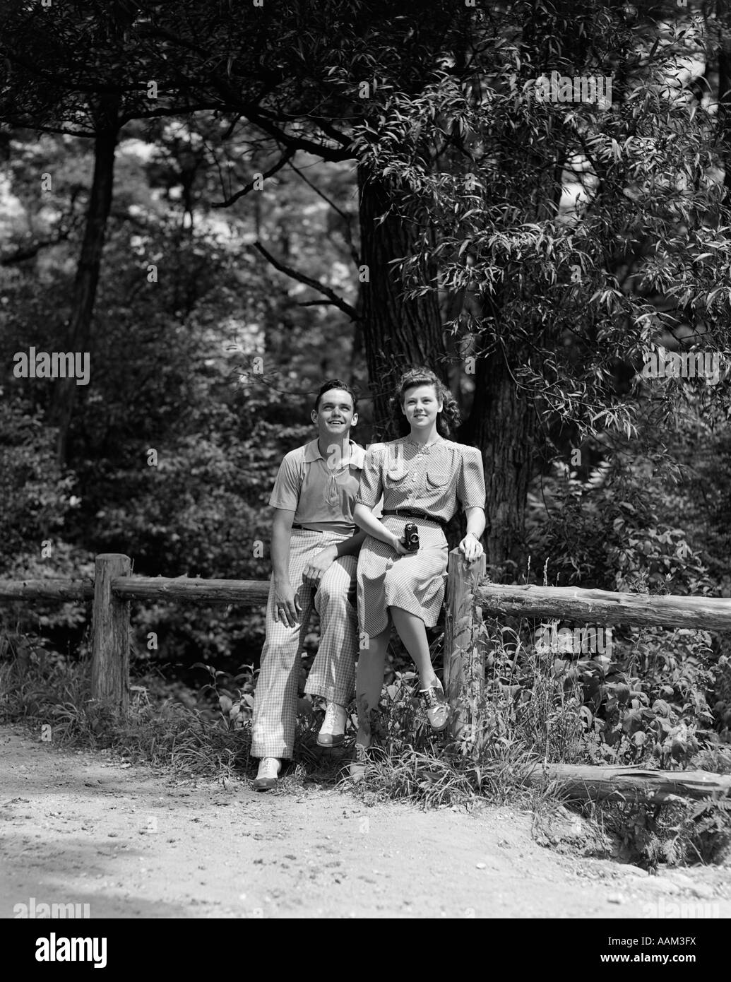 1940ER JAHRE TEEN PAAR SITZT AM ZAUN IM WALD AUF DER SUCHE, WEIBLICH, DIE KAMERA ZU HALTEN Stockfoto