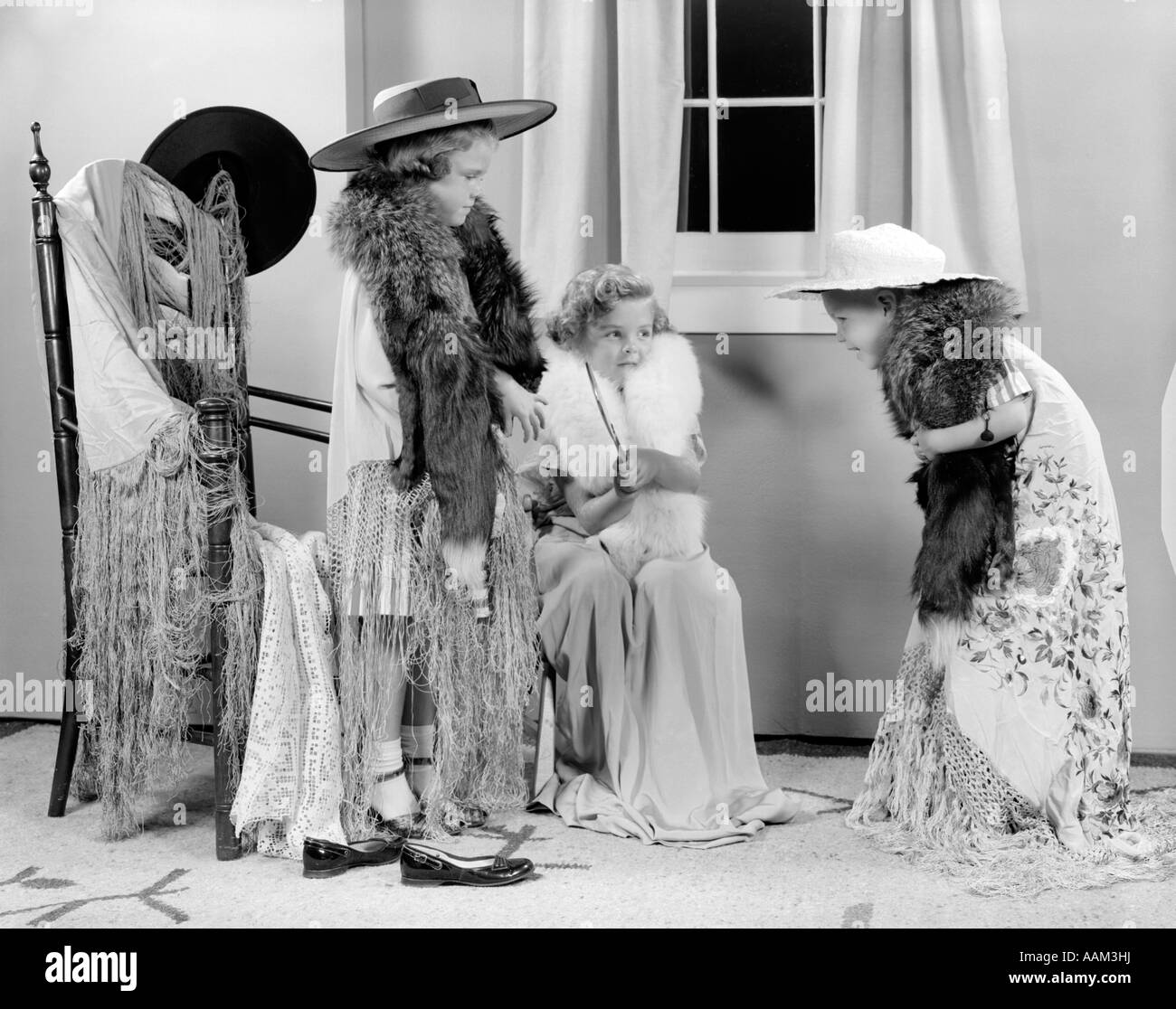 1950ER JAHREN DREI KINDER ALLE GEKLEIDET WIE ERWACHSENE FRAUEN IN SCHALS PELZKRAGEN HÜTE VOR VERHANGENEN FENSTER SPIELEN SOMMER Stockfoto