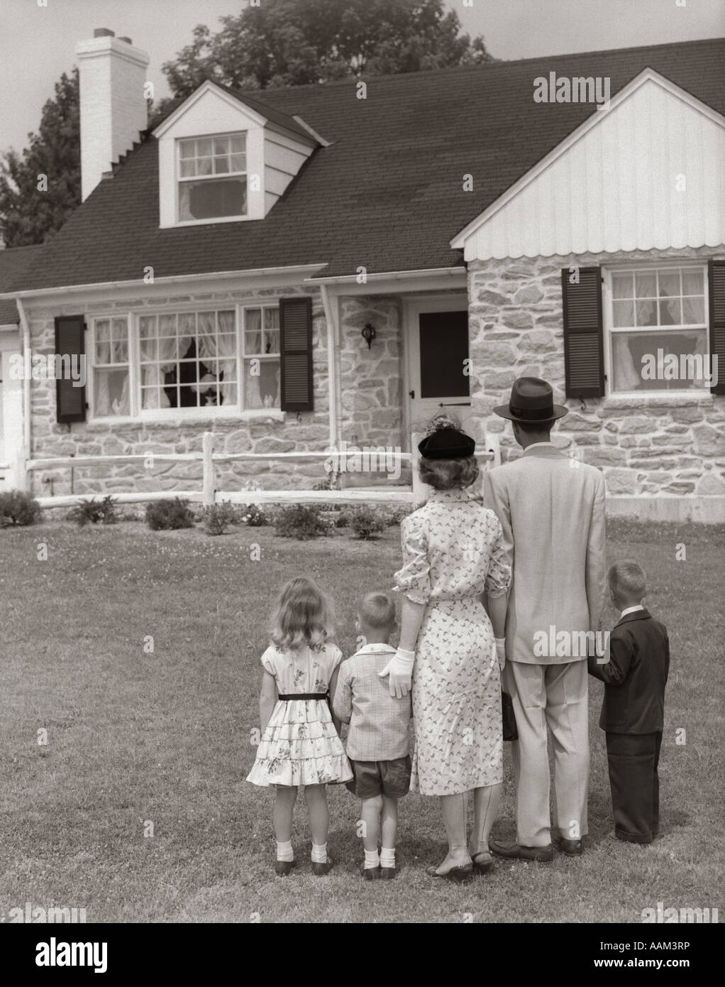 1950ER JAHRE 5-KÖPFIGE FAMILIE MIT DEM RÜCKEN ZUR KAMERA AUF RASEN FELDSTEINEN HAUS BETRACHTEN Stockfoto