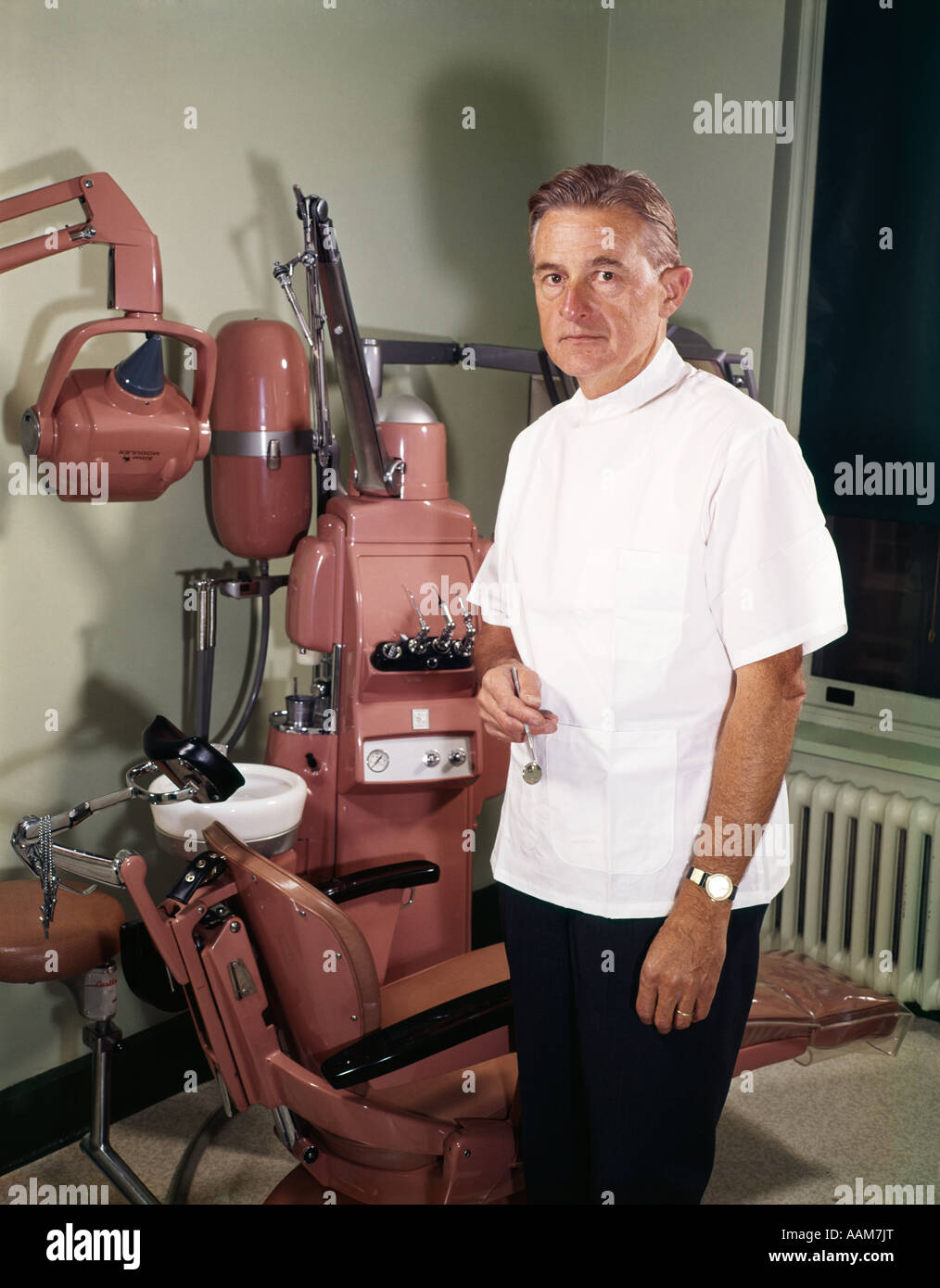 MITTE DER 1960ER JAHRE IM ALTER VON ZAHNARZT TRAGEN WEIßE JACKE STANDBY STUHL IM BEHANDLUNGSRAUM MIT ZAHNBOHRER UND WASCHBECKEN BLICK IN DIE KAMERA Stockfoto