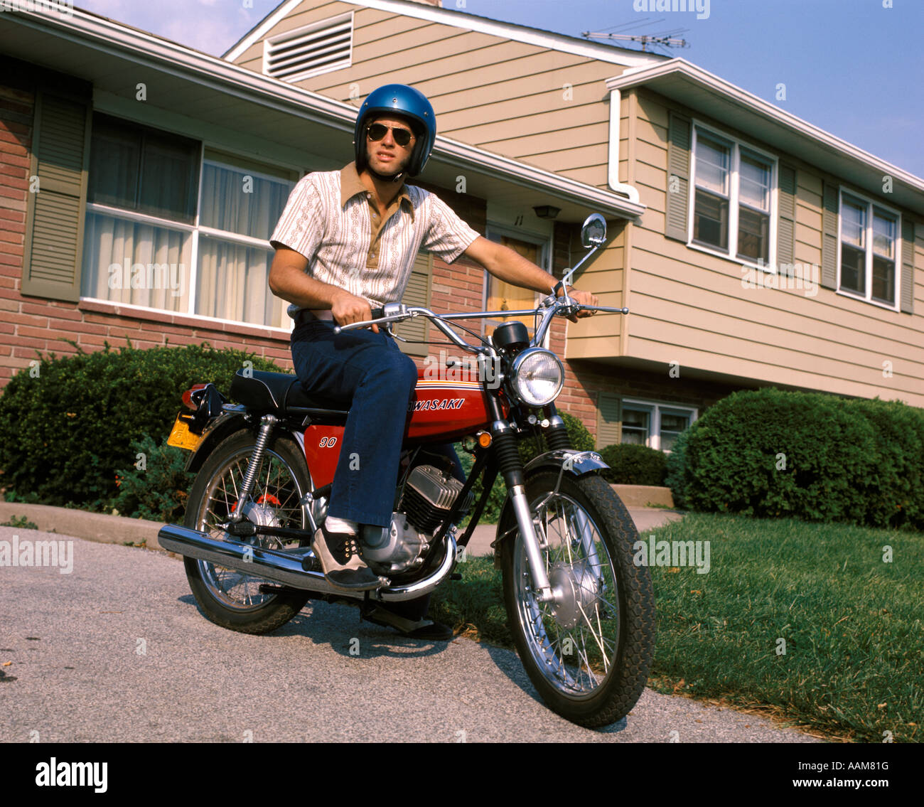 1970 1970S JUNGER MANN BLAUEN HELM SITZT AUF DEM MOTORRAD IN EINFAHRT  VORSTADTHAUS BIKE MOTORRÄDER MÄNNER Stockfotografie - Alamy