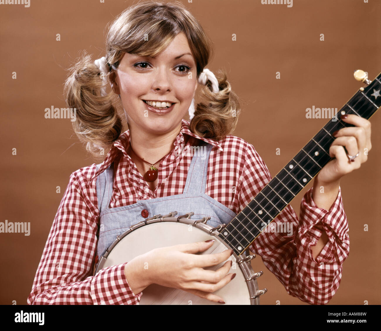 1970ER JAHRE JUNGE FRAU IN KAROHEMD UND OVERALLS MIT HAARE IM PFERDESCHWANZ SPIELT BANJO KLIMPERN RUBE BUMPKIN LAND Stockfoto