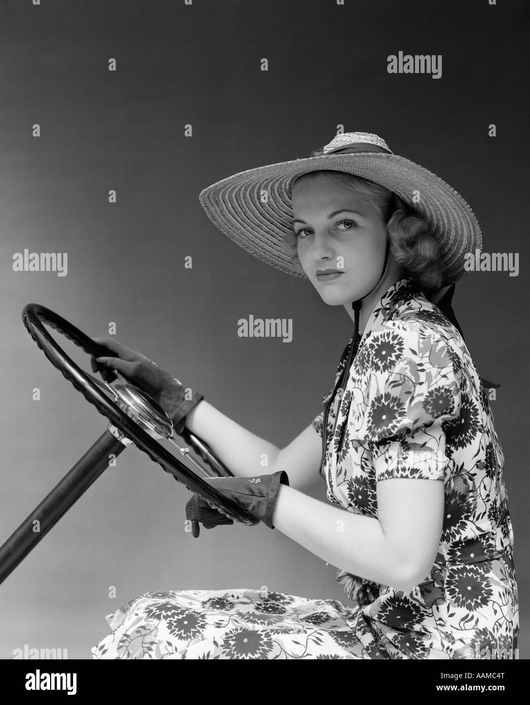 1930S 1940S AUTO-FRAU TRAGEN STROHHUT SONNE KLEID FAHREN HANDSCHUHE HALTEN MOBILE LENKRAD BLICK IN DIE KAMERA Stockfoto