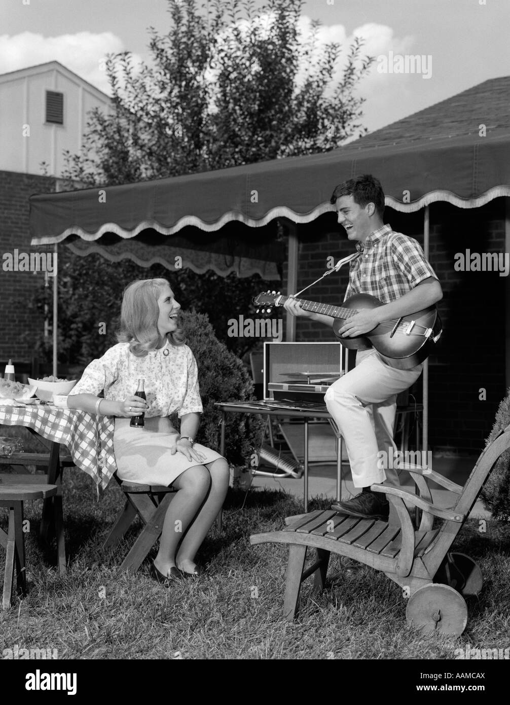 1960ER JAHRE TEENAGER-PAAR BOY SPIELEN GITARRE WOMAN SITTING ON GARTENMÖBEL MIT SOFTDRINK FLASCHE DRAUßEN IM HINTERHOF Stockfoto