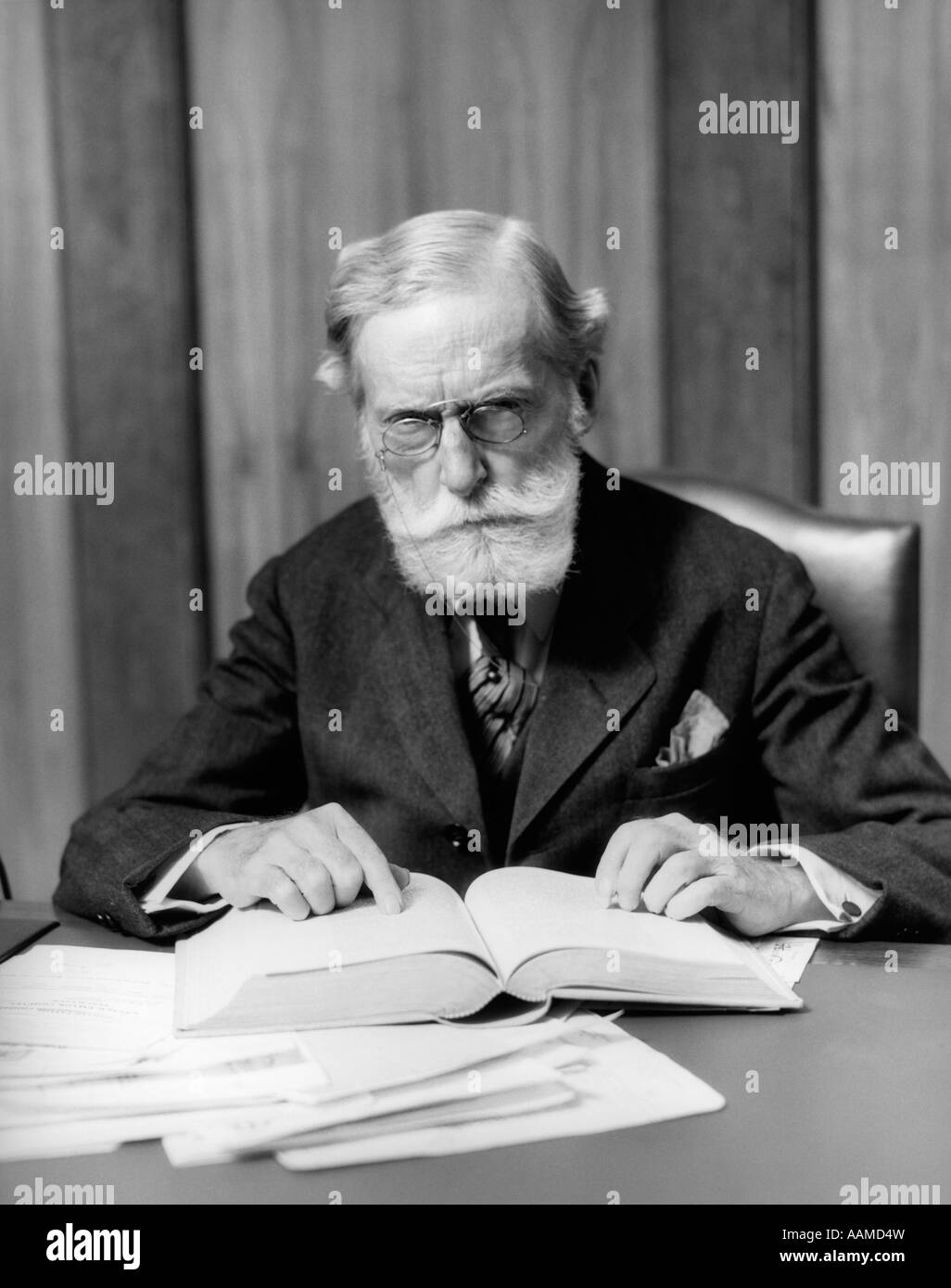 1930ER JAHRE ÄLTEREN MANN IN GLÄSERN SITZEN AM SCHREIBTISCH-LESEBUCH Stockfoto