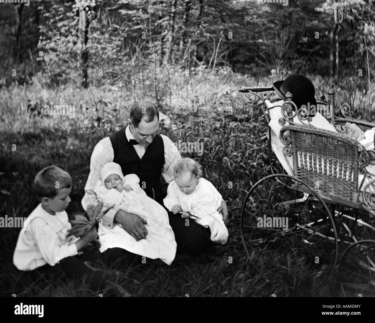 1890S 1900S WENDE DES JAHRHUNDERTS VATER SITZEN IM WALD UND HAT DREI KINDER BABY CARRIAGE AUS ZUR SEITE Stockfoto