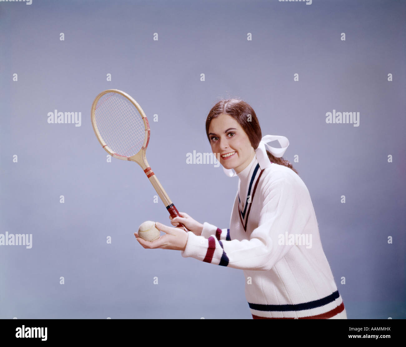 1960ER JAHREN FRAU TENNIS SPIELER HOLDING BALL UND SCHLÄGER BEREIT, PULLOVER LÄCHELND ZU DIENEN Stockfoto