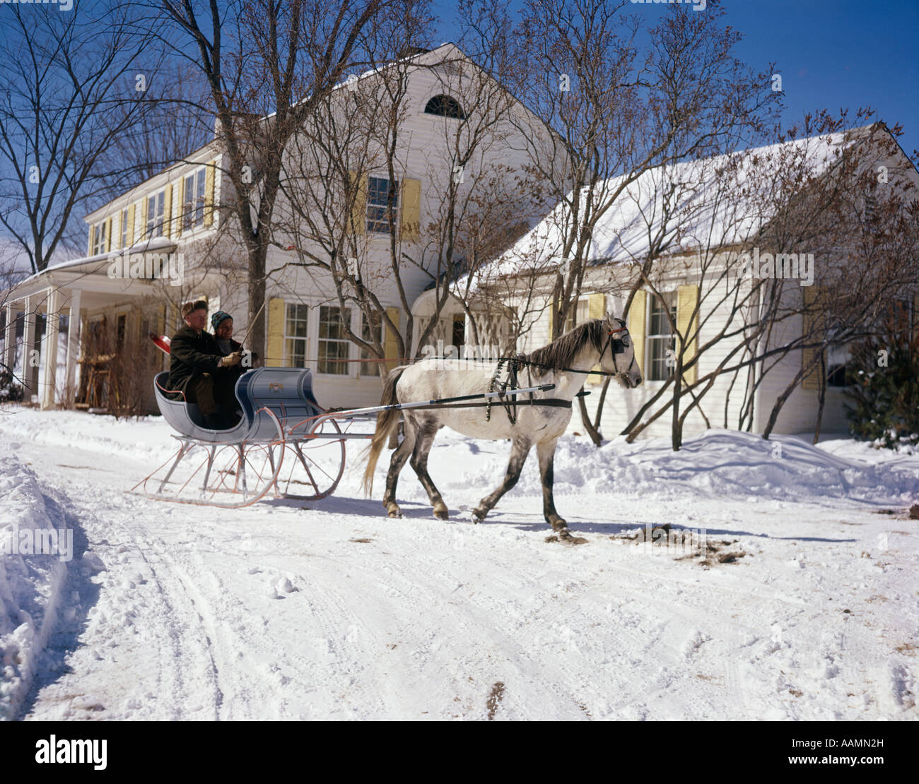 1960ER JAHRE SCHNEE 2 MÄNNER IN EIN WEIßES PFERD GEZEICHNETEN OFFENEN SCHLITTEN VON HAUS LANDGASTHOF RETRO VINTAGE WINTER VERMONT Stockfoto