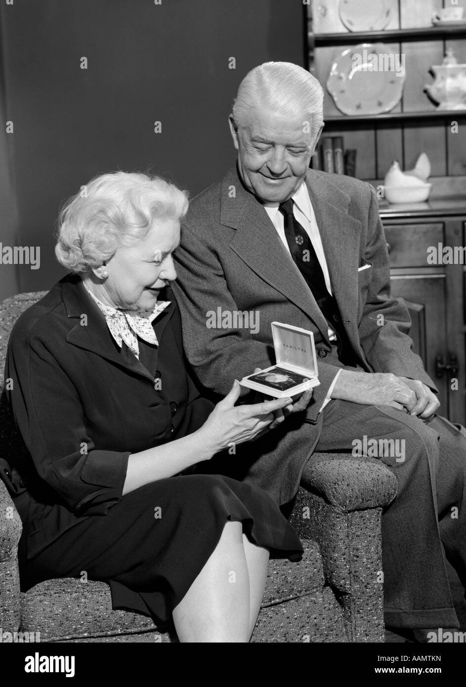 1950ER JAHRE ÄLTERE FRAU SITZT IM SESSEL ERÖFFNUNG SCHMUCK GESCHENK-BOX MIT EHEMANN AUF ARM DER STUHL SITZEND Stockfoto