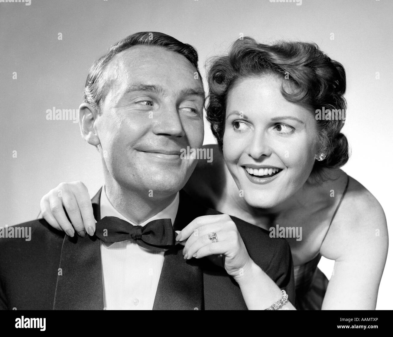 1950S 1960S PAAR LÄCHELND IN FORMALEN ABEND KLEIDUNG FRAU ZIEHEN AM MANNES FLIEGE GEKLEIDET Stockfoto