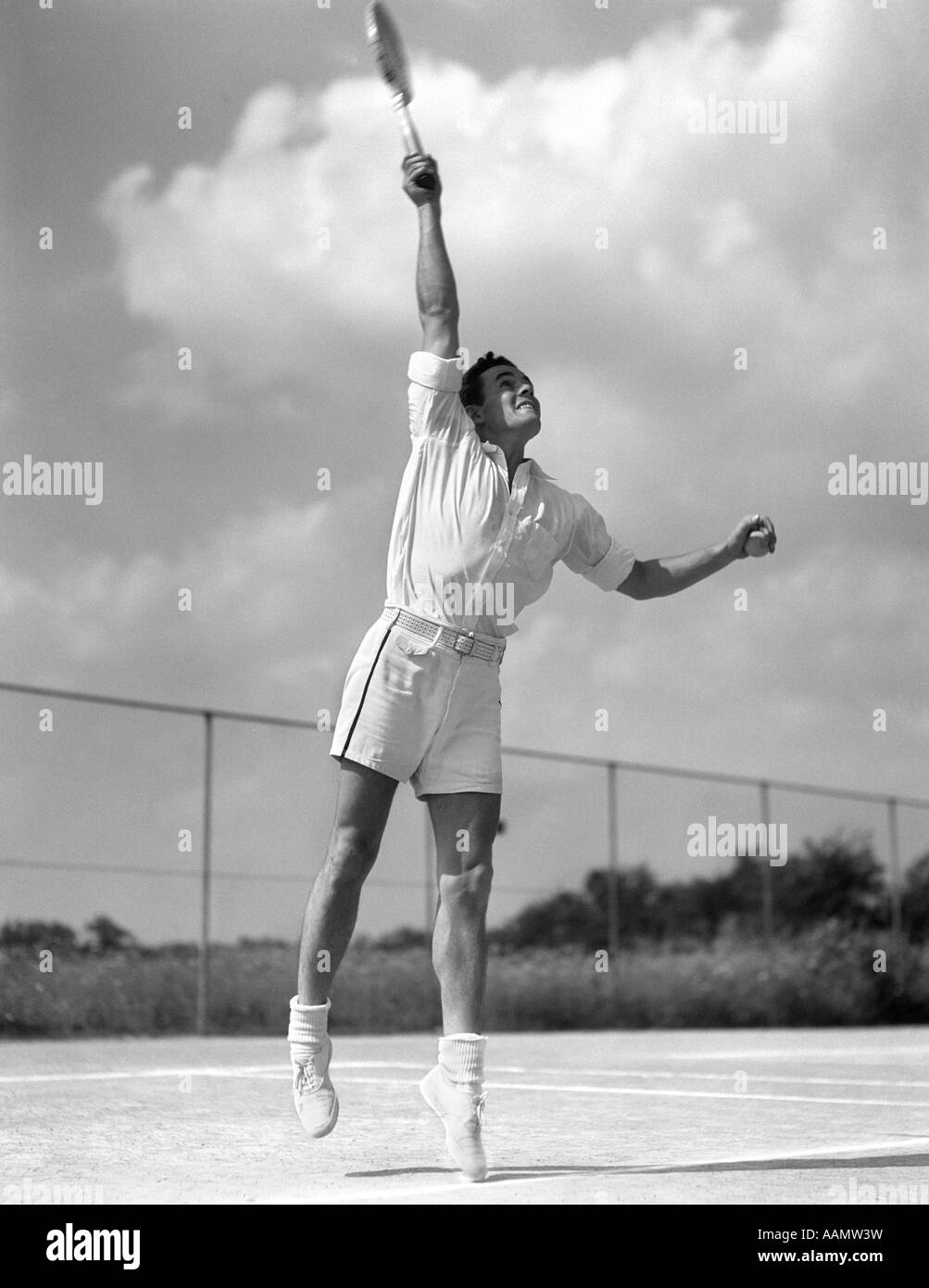 1930ER JAHRE MANN IN DER MITTE DIENEN SPIELEN TENNIS SCHWINGENDEN TENNISSCHLÄGER UM BALL SPRINGEN Stockfoto