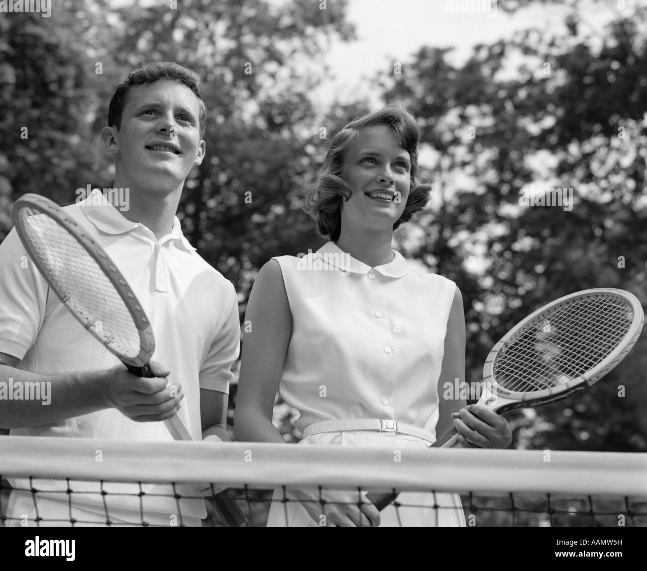 1960ER JAHREN LÄCHELND PORTRAIT PAAR HINTER NET HOLDING TENNISSCHLÄGER Stockfoto