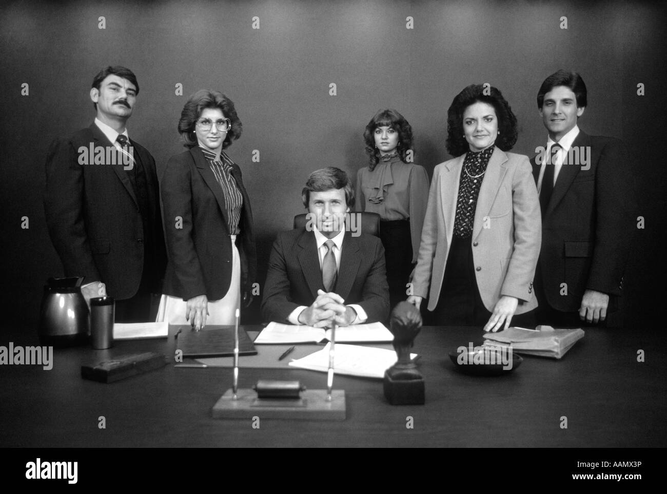 1980ER JAHRE GRUPPENPORTRÄT-OFFICE-MITARBEITER STEHEN HINTER SCHREIBTISCH RUND UM CHEF MIT SCHWEREN AUSDRÜCKE Stockfoto