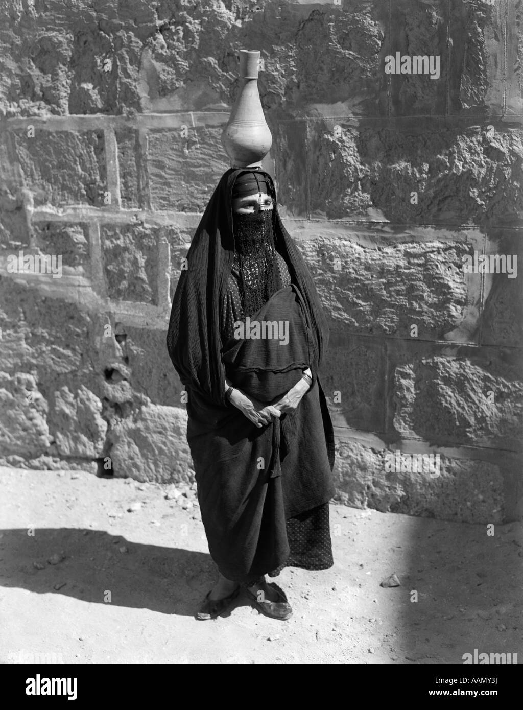 1920S 1930S ÄGYPTISCHE FRAU MIT KRUG AUF KOPF BALANCIERT TRAGEN TRADITIONELLE ARABISCHE MUSLIMISCHE GEWAND GESICHT VERSCHLEIERT BURKA Stockfoto