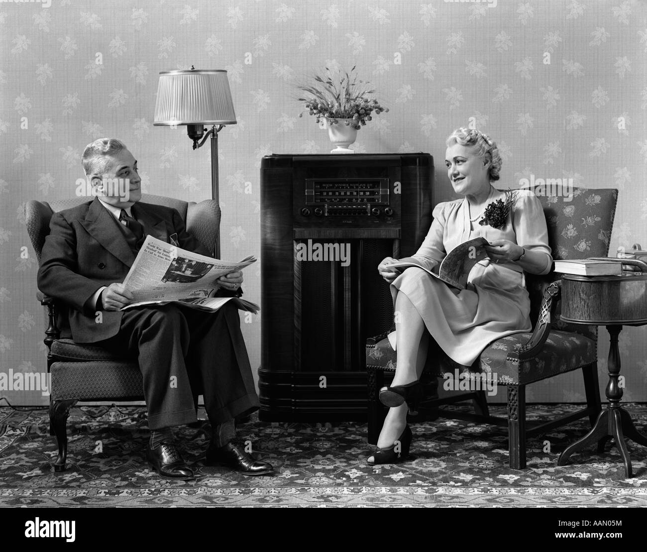 1940ER JAHRE ÄLTERES PAAR SITZEN IM WOHNZIMMER LESEN ZEITUNGEN UND ZEITSCHRIFTEN, RADIO HÖREN Stockfoto