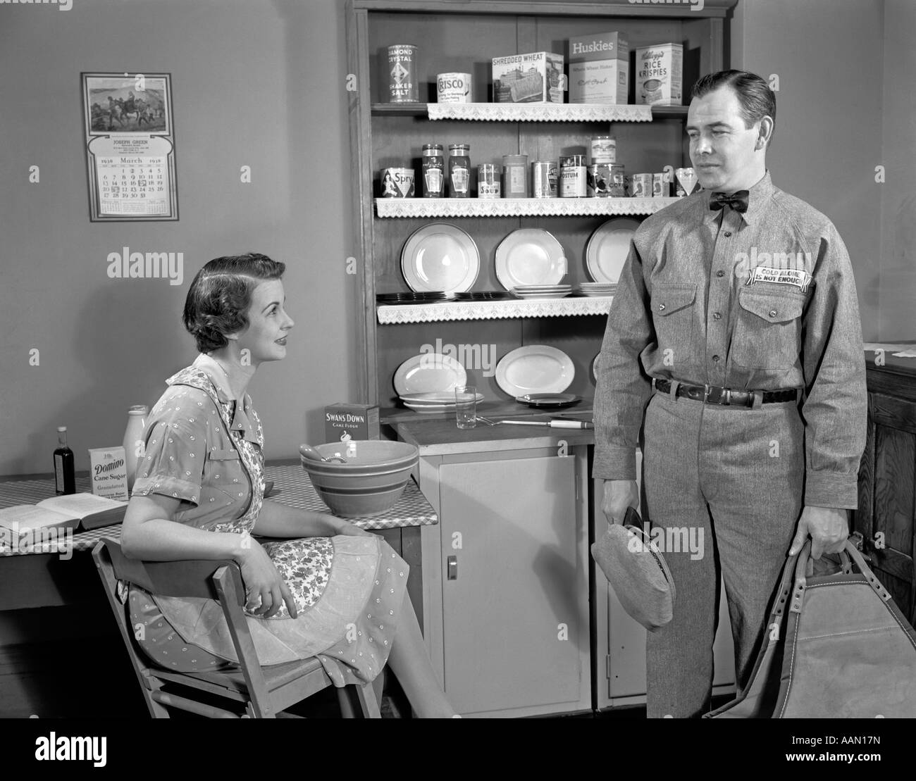 1940ER JAHREN FRAU SCHÜRZE SITZT AM KÜCHENTISCH, IM GESPRÄCH MIT MECHANIKER ODER MANN IN UNIFORM HAUSFRAU ESSEN KARTONS IN REGALEN Stockfoto