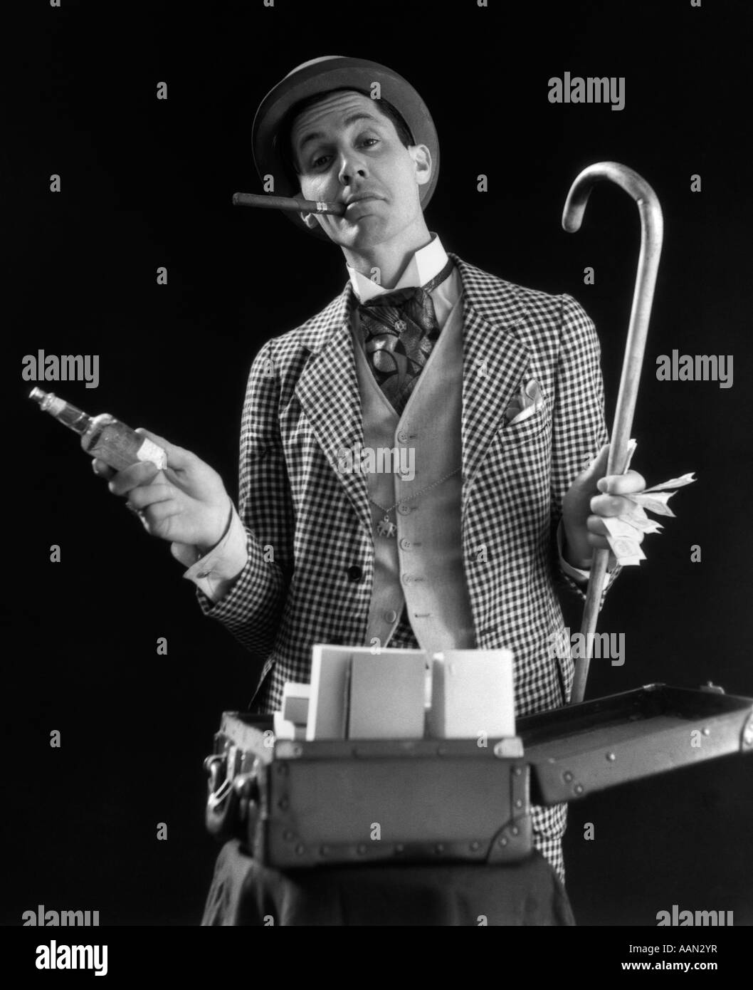 1930ER JAHREN BARKER KARIERTEN ANZUG ZIGARRE IM MUND HOLDING CANE GELD FLASCHE TONIC OFFEN KOFFERABLAGE, BLICK IN DIE KAMERA Stockfoto