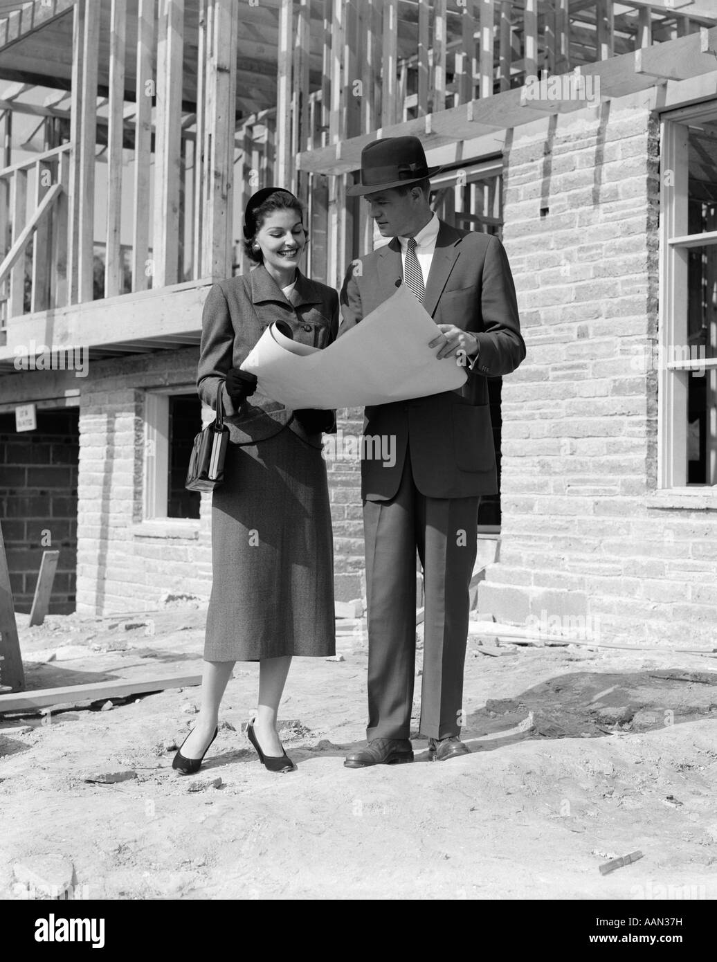 1950ER JAHREN LÄCHELND PAAR BLICK AUF GEBÄUDE PLANT STAND VOR UNFERTIGE HAUS Stockfoto