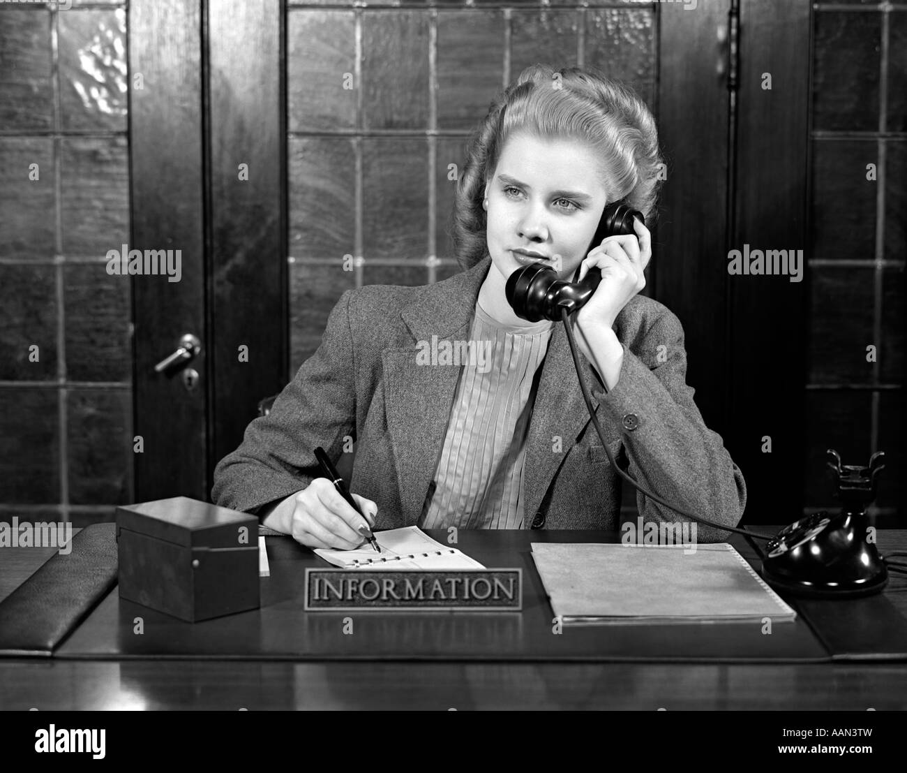 1940ER JAHRE JUNGE FRAU SITZT AM INFORMATION DESK SEKRETÄR GESPRÄCH AM TELEFON SCHREIBEN Stockfoto