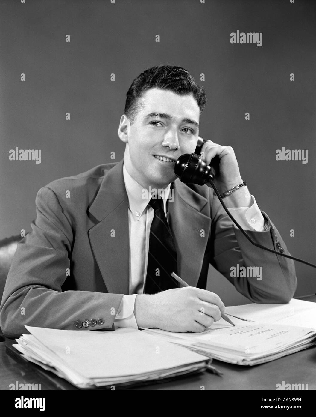 1950ER JAHRE JUNGEN GESCHÄFTSMANN GESPRÄCH AM TELEFON SITZEN AM SCHREIBTISCH SCHREIBEN Stockfoto