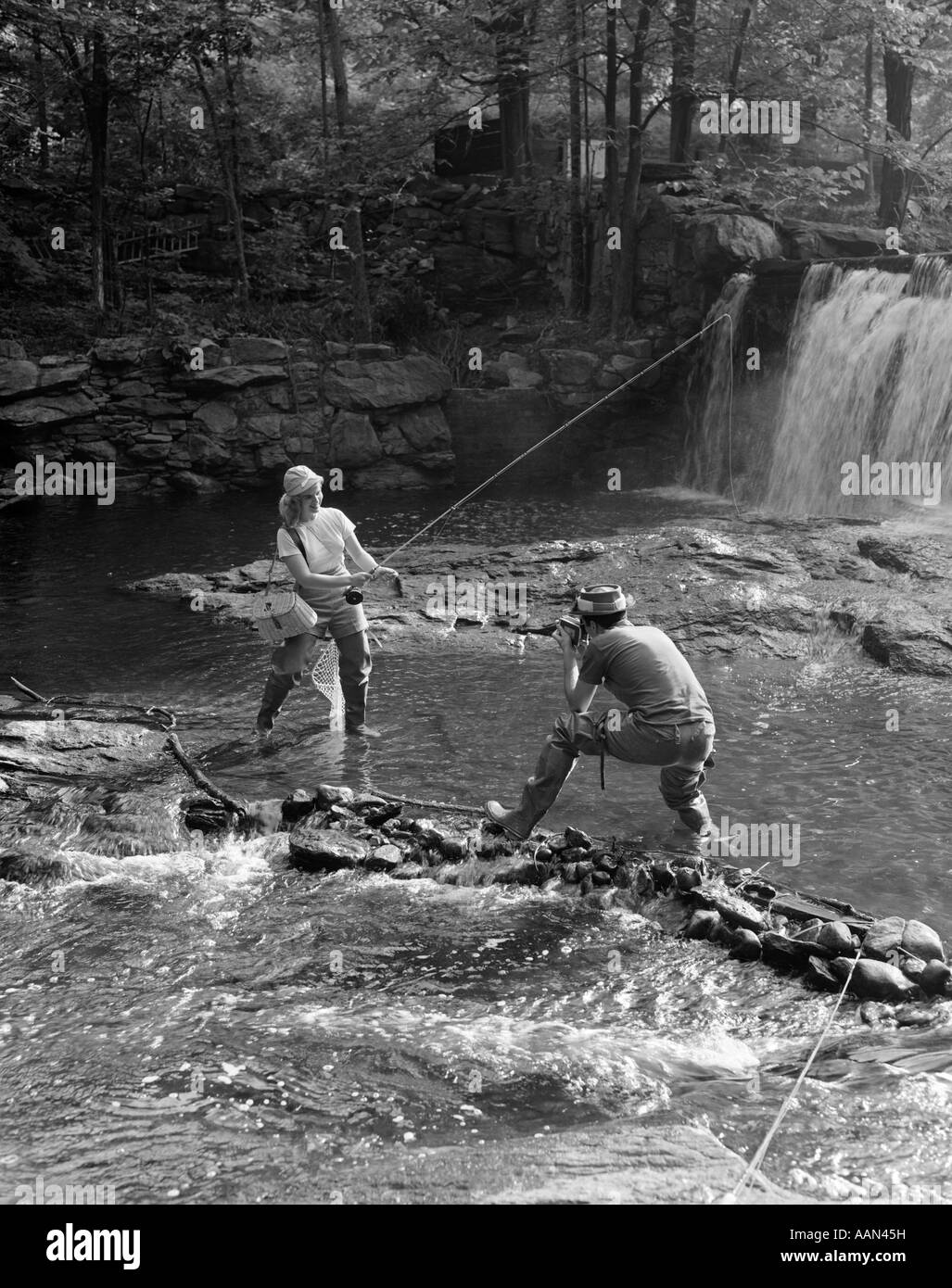 1950ER JAHREN PAAR MANN NEHMEN FOTO VON FRAU FLIEGENFISCHEN IN BACH MIT WASSERFALL Stockfoto