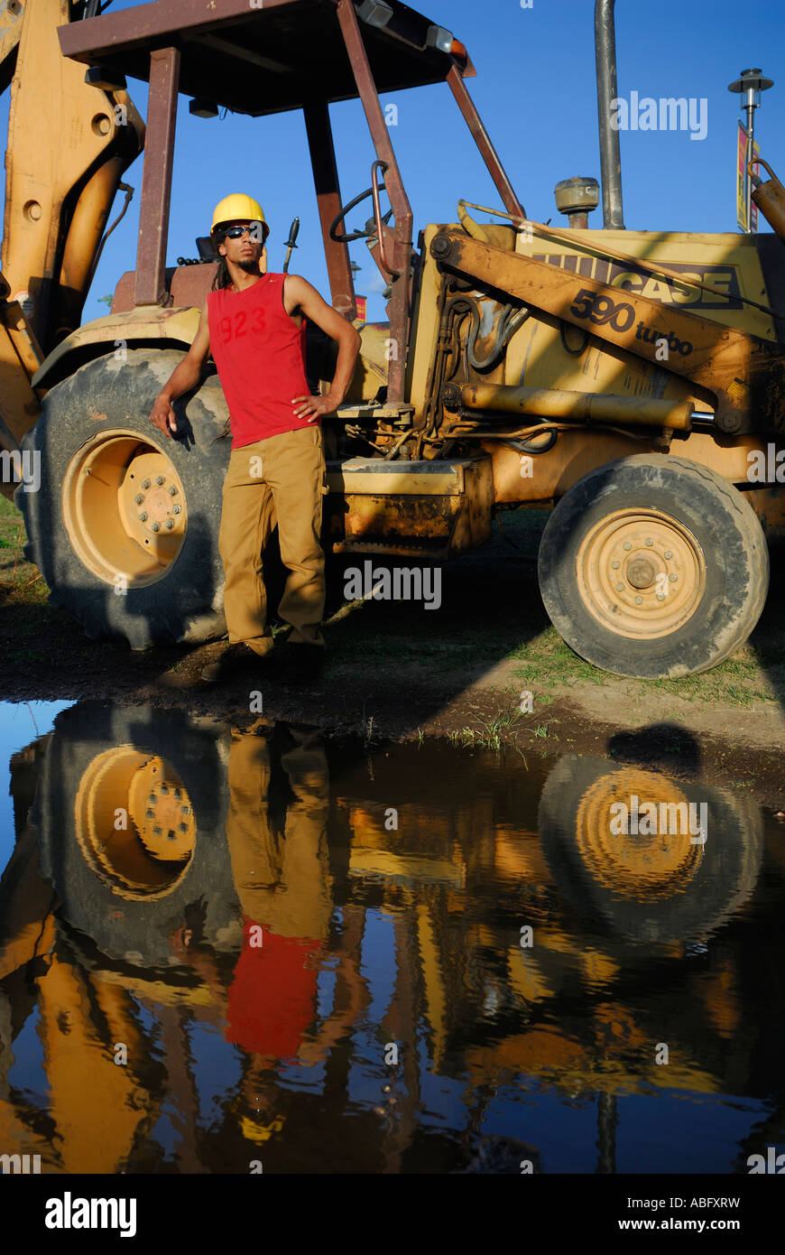 Schwarzen männlichen Bauarbeiter in Schutzhelm stützte sich auf Baggerlader mit Spiegelbild im Wasser Pfütze Stockfoto