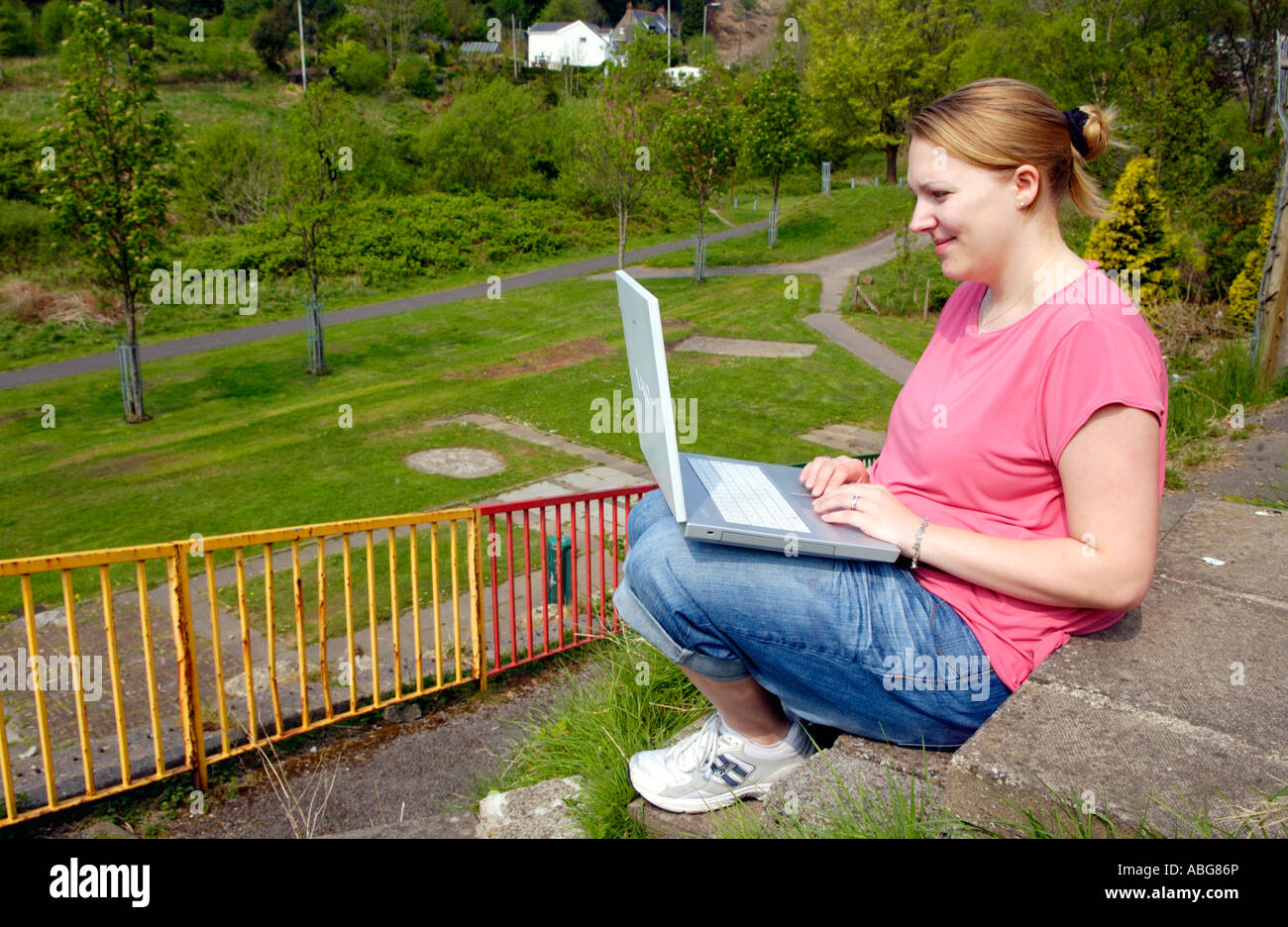 Junge Frau saß draußen im öffentlichen Park mit Sony Vaio Wireless-Laptop in Jeans rosa t-Shirt gekleidet und Trainer UK Stockfoto