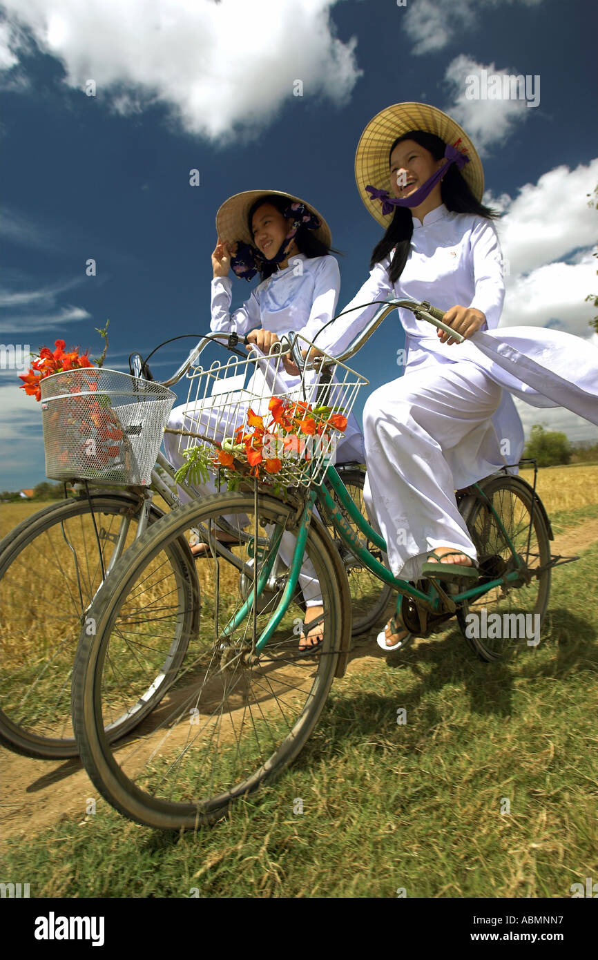 Zwei junge Frauen in der konischen Hüten und traditionelle Kostüme fahren Fahrräder durch Reisfelder in der Nähe von Phan Thiet Vietnam Stockfoto