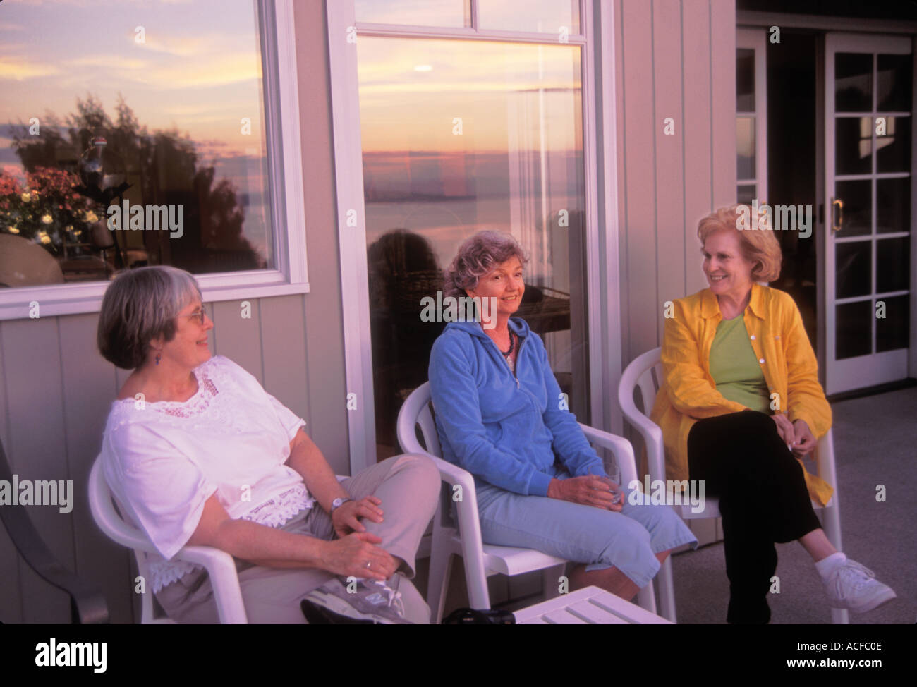 Drei Frauen in Führungspositionen sitzen friedlich lächelnd und sprechen auf der Veranda des Hauses bei Sonnenuntergang im Sommer mit Reflexionen im Fenster Stockfoto