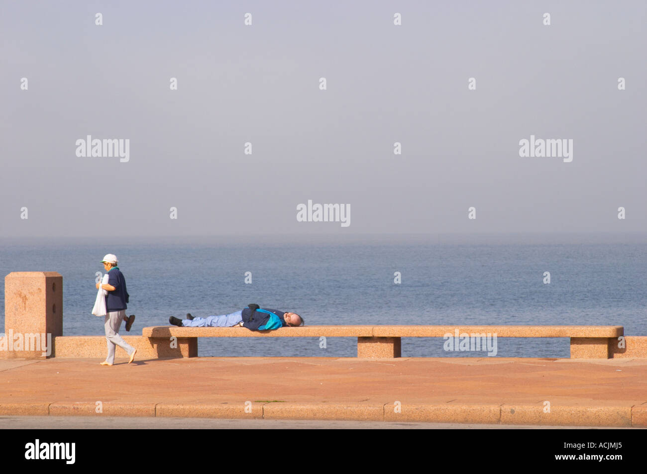 Eine Frau zu Fuß auf dem Bürgersteig mit einer Thermoskanne heißes Wasser mit Mate Kräutertee und ein Mann auf einer Bank schlafen, auf dem Spaziergang am Flussufer am Meer entlang des Flusses liegend, Rio De La Plata Ramblas Sur, Gran Bretagna und Republica Argentina Montevideo, Uruguay, Südamerika Stockfoto