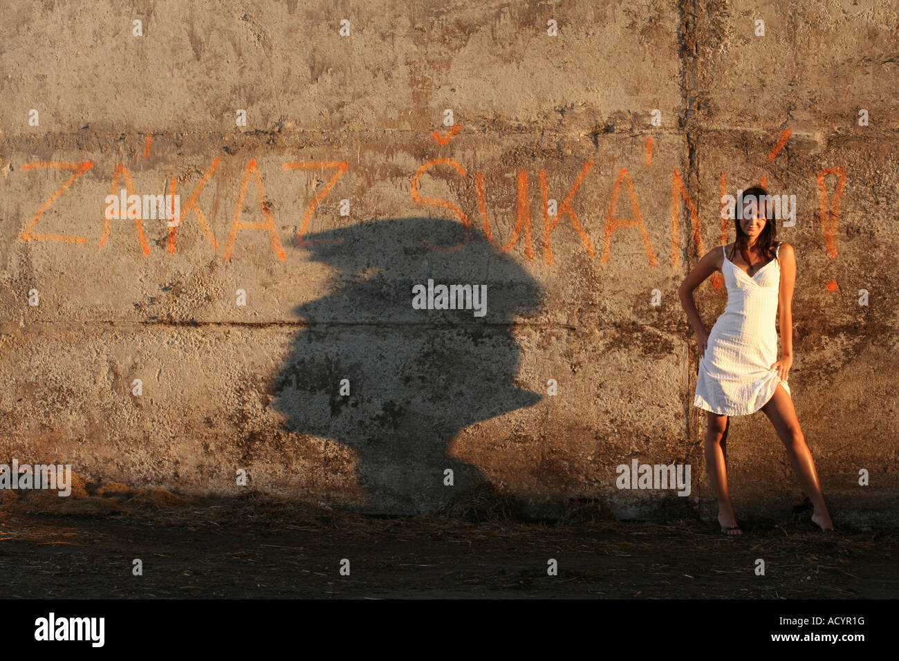 Eine Frau in einem weißen Kleid, die bei Sonnenuntergang neben einer mit Graffiti bedeckten Betonmauer posiert und einen dramatischen Schatten wirft, in einer städtischen Umgebung in Tschechien Stockfoto