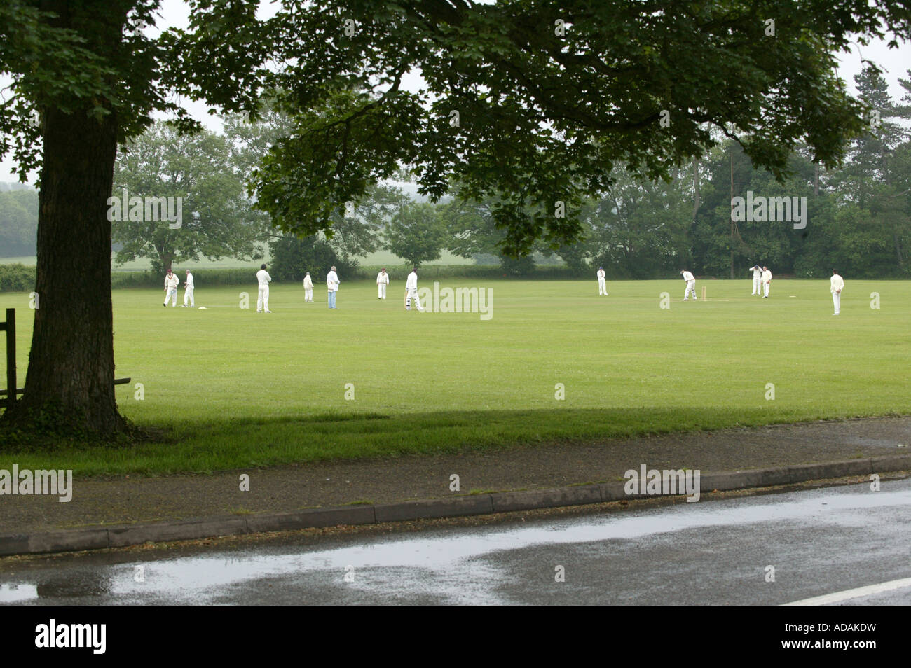 Ein ländliches Dorf Spiel Cricket in der englischen Landschaft mit Regen auf der Straße im Vordergrund Stockfoto