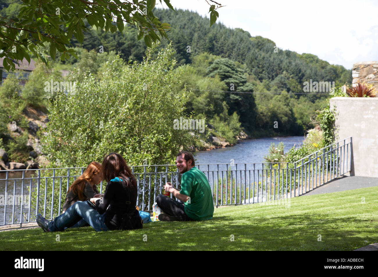 drei ausländische Touristen machen eine spontanes Picknick, sitzen auf Rasen vor Zaun mit Blick auf Fluss im Tal von avoca Stockfoto