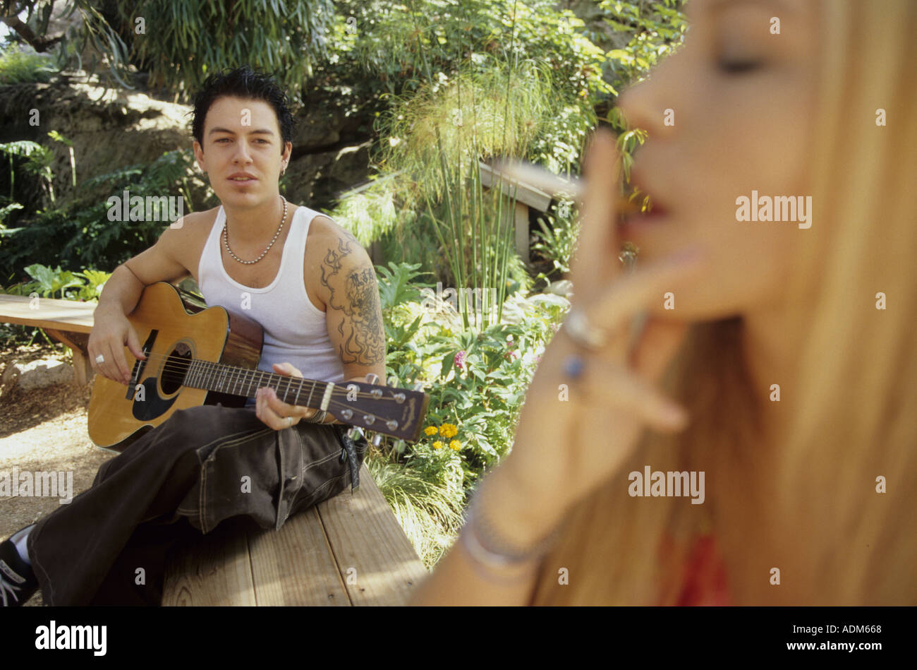 Ein Junger Mann Mit Tattoos Spielt Seine Gitarre Im Hintergrund Wahrend Das Junge Madchen Eine Zigarette Im Vordergrund Raucht Stockfotografie Alamy