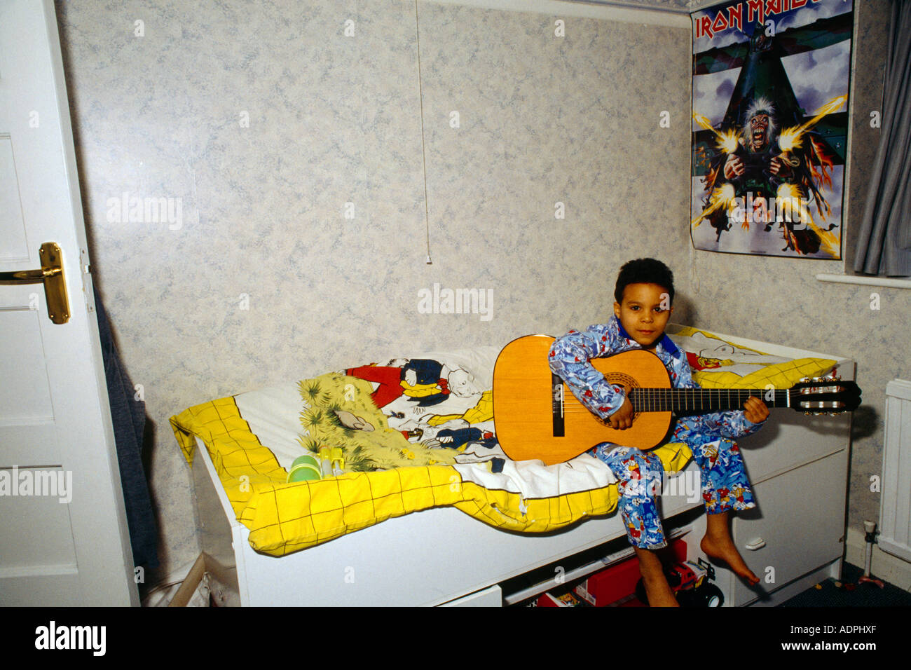Afro Karibik junge im Baumwolle Fußball Pyjama spielt akustische Gitarre auf Bett sitzend mit Rupert Bär-Baumwollbezug Stockfoto