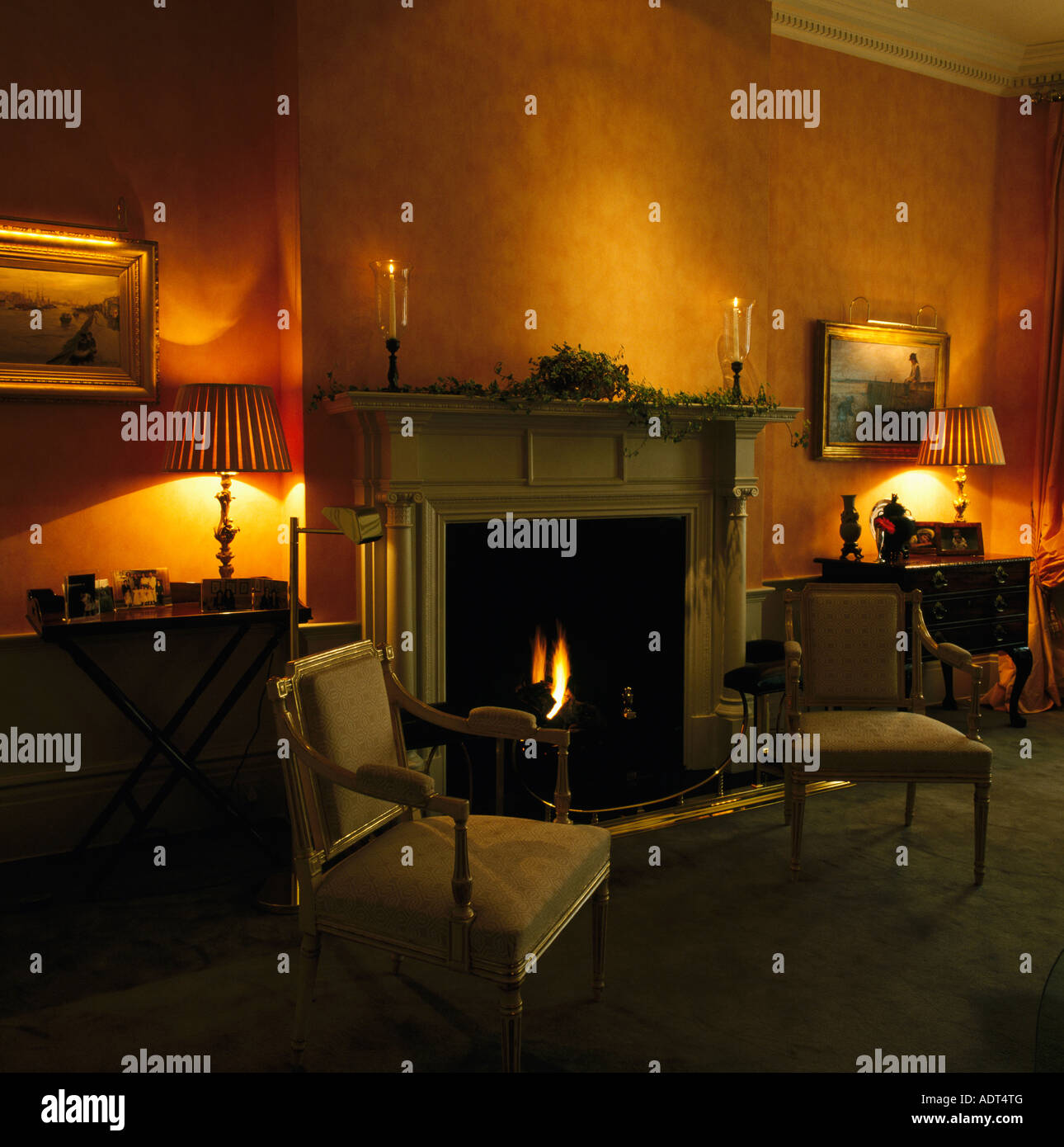 Stühle und brennenden Lampen auf beiden Seiten des Kamins mit Feuer angezündet in abgedunkelten Stadthaus-Wohnzimmer Stockfoto