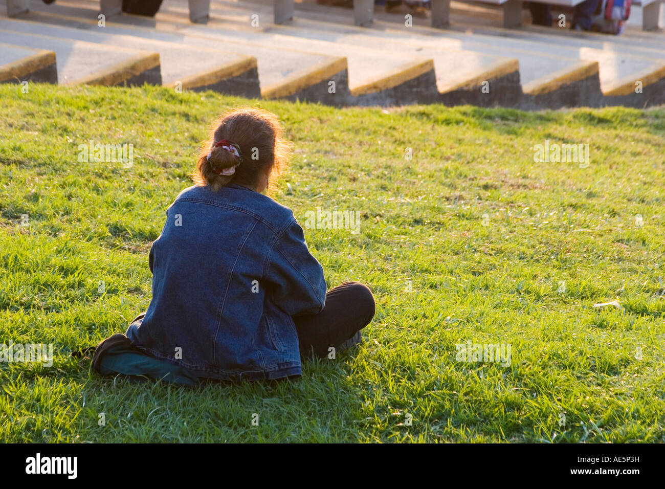 Zurück von einer rothaarigen Frau sitzt auf einem Rasen Hügel in einem Stadion beobachten ein lokales Ereignis in der Nachmittagssonne Stockfoto