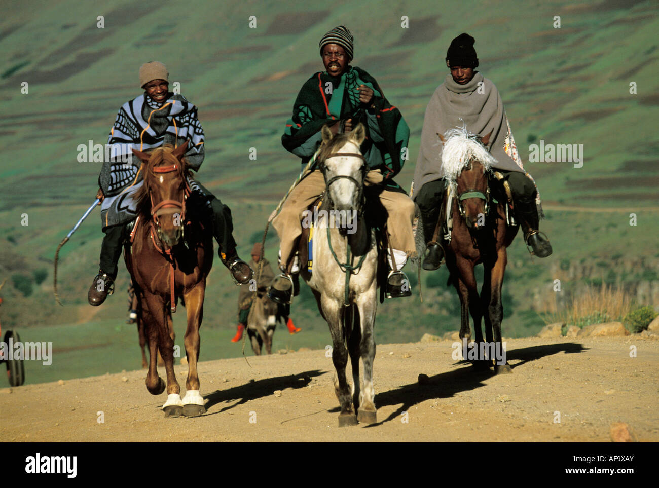 Drei Männer mit Decken eingewickelt um sie herum reiten auf einem Schotterweg in einer bergigen Gegend von Lesotho Stockfoto