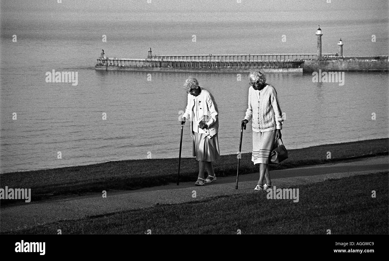 Zwei alte Damen mit Stöcken zu Fuß entlang der Klippe bei Whitby North Yorkshire mit den Hafen-Landungsbrücken in der backgr Stockfoto