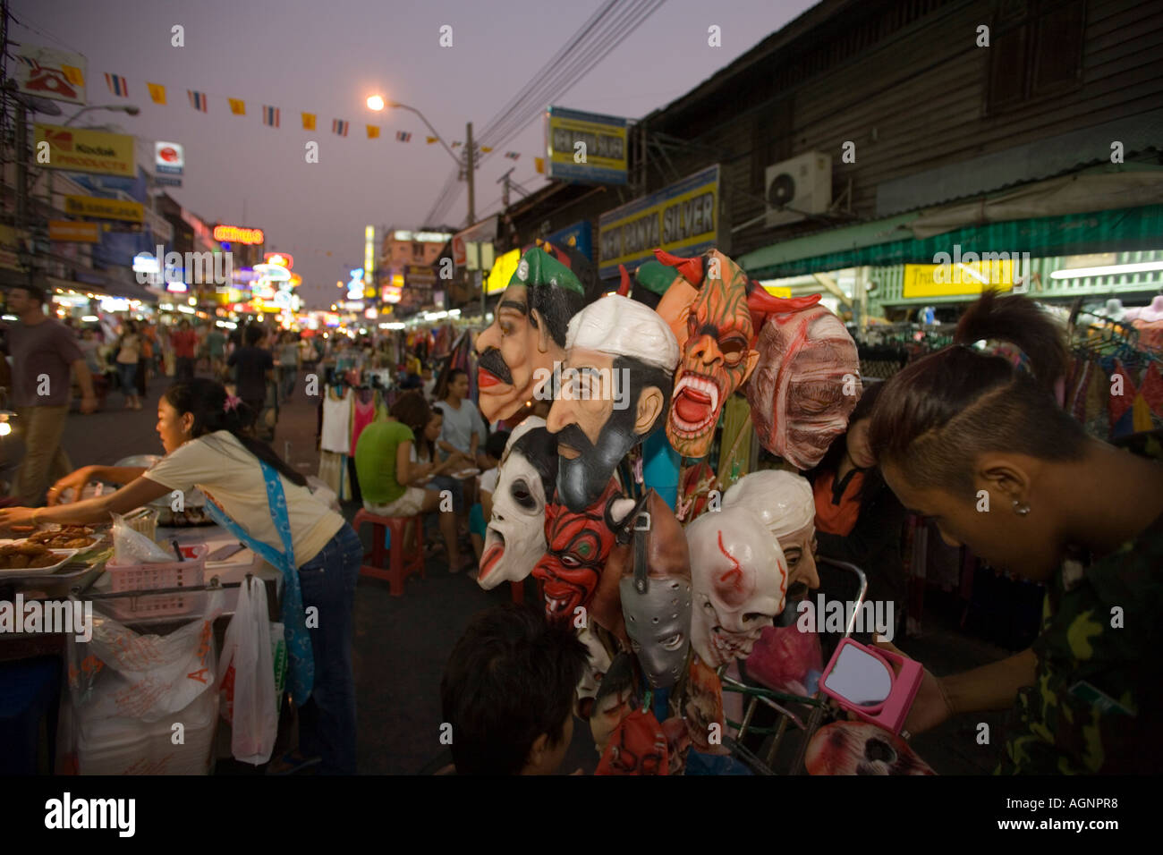 Menschen beim Einkaufen bei Th Khao San Road in den Abend ein Ständer mit Masken im Vordergrund Banglamphu Bangkok Thailand Stockfoto