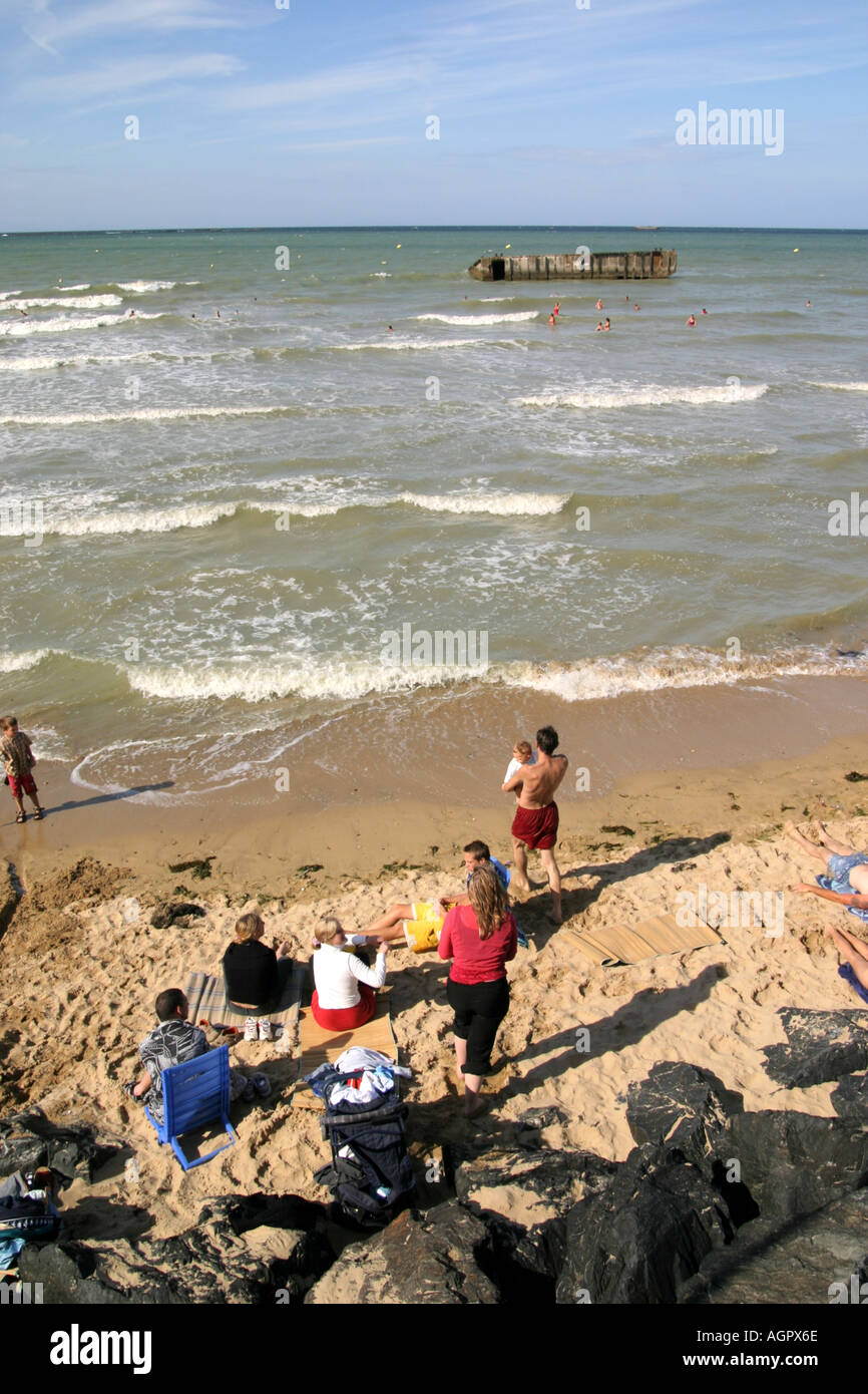 Badegäste am Strand von Arromanches-Les-Bains in Normandie Frankreich mit Trümmern der alten Häfen Maulbeere im Hintergrund Stockfoto