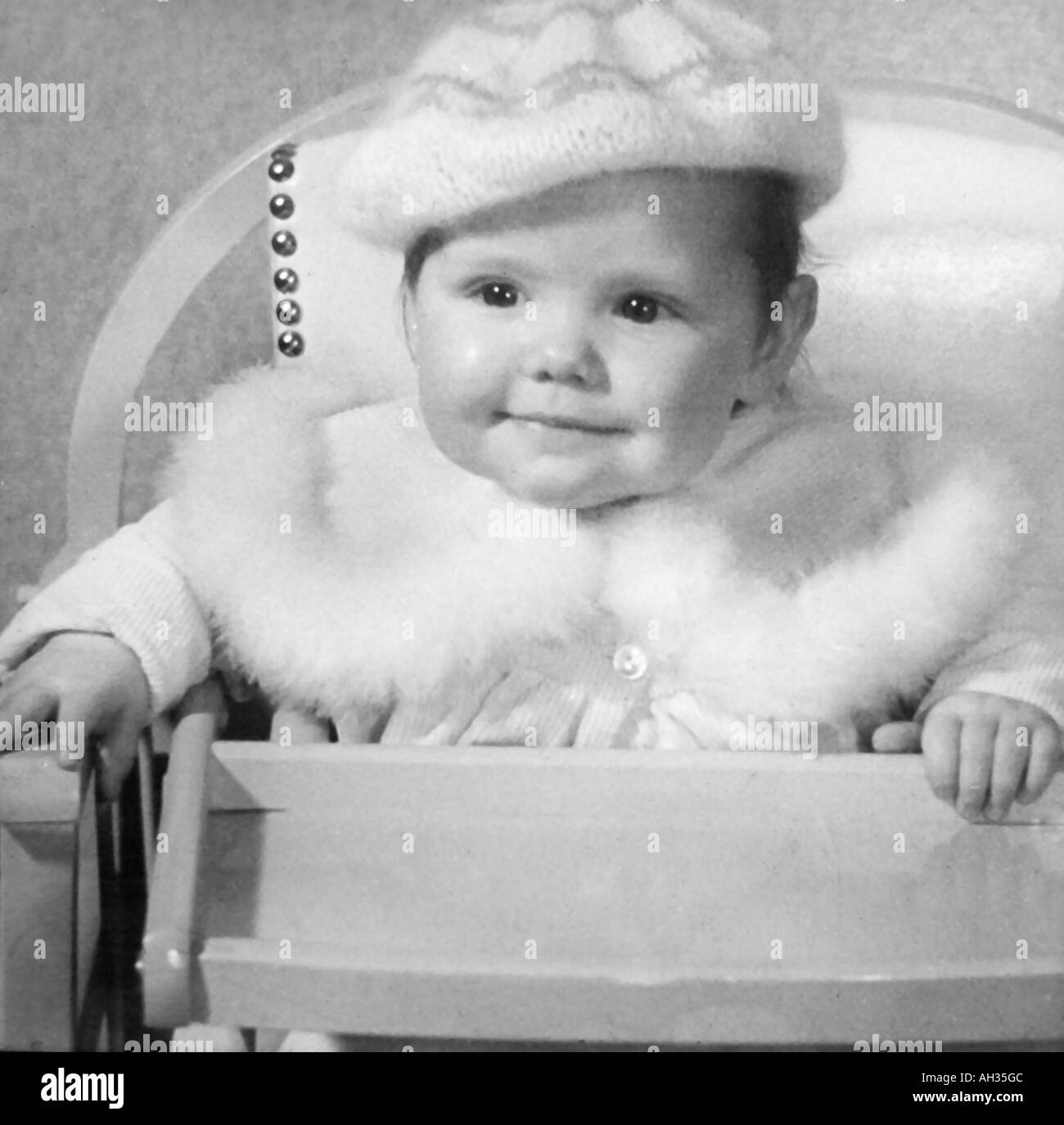 ALTE VINTAGE SCHWARZ / WEIß FOTO SNAP SHOT BABY MÄDCHEN TRAGEN GETRIMMTE PELZ UND WOLLE MÜTZE IM HOCHSTUHL SITZEN Stockfoto