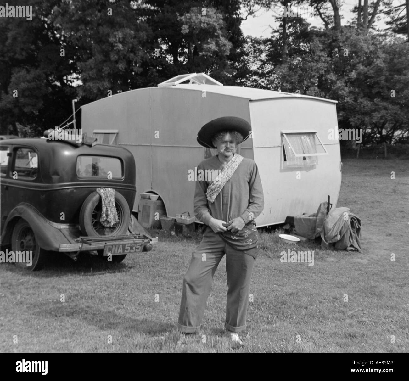 ALTE VINTAGE SCHWARZ / WEIß FAMILIE SNAPSHOT FOTO MANN GEKLEIDET IN COWBOY KOSTÜM STEHEN IM VORDERGRUND DES ALTEN AUTO UND CARAVA Stockfoto