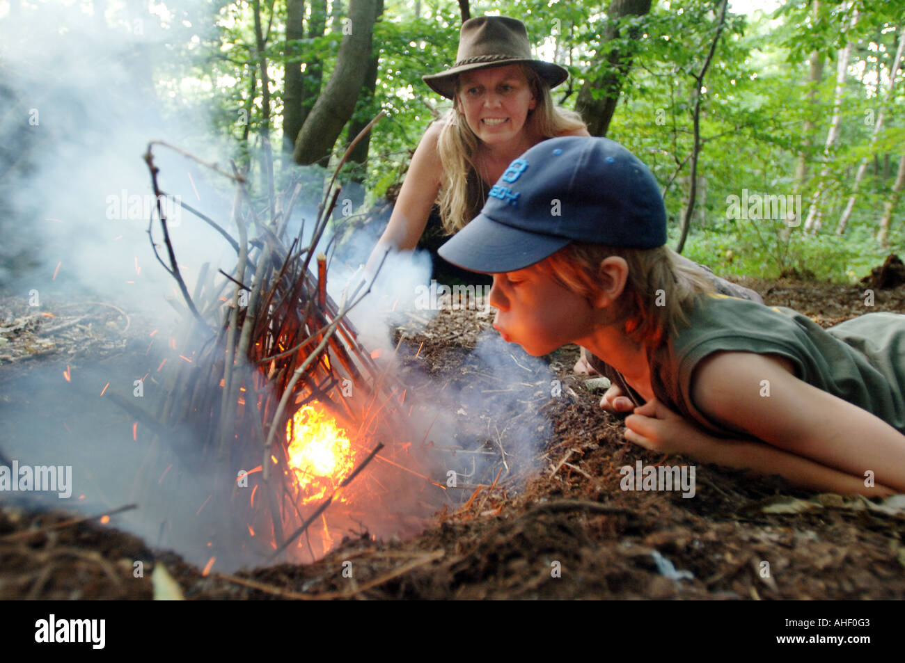 Kleiner Junge in Baseball-Mütze weht einen Lagerfeuer in das Leben in einem Waldgebiet clearing als Lehrerin Frau blickt auf zustimmend Stockfoto