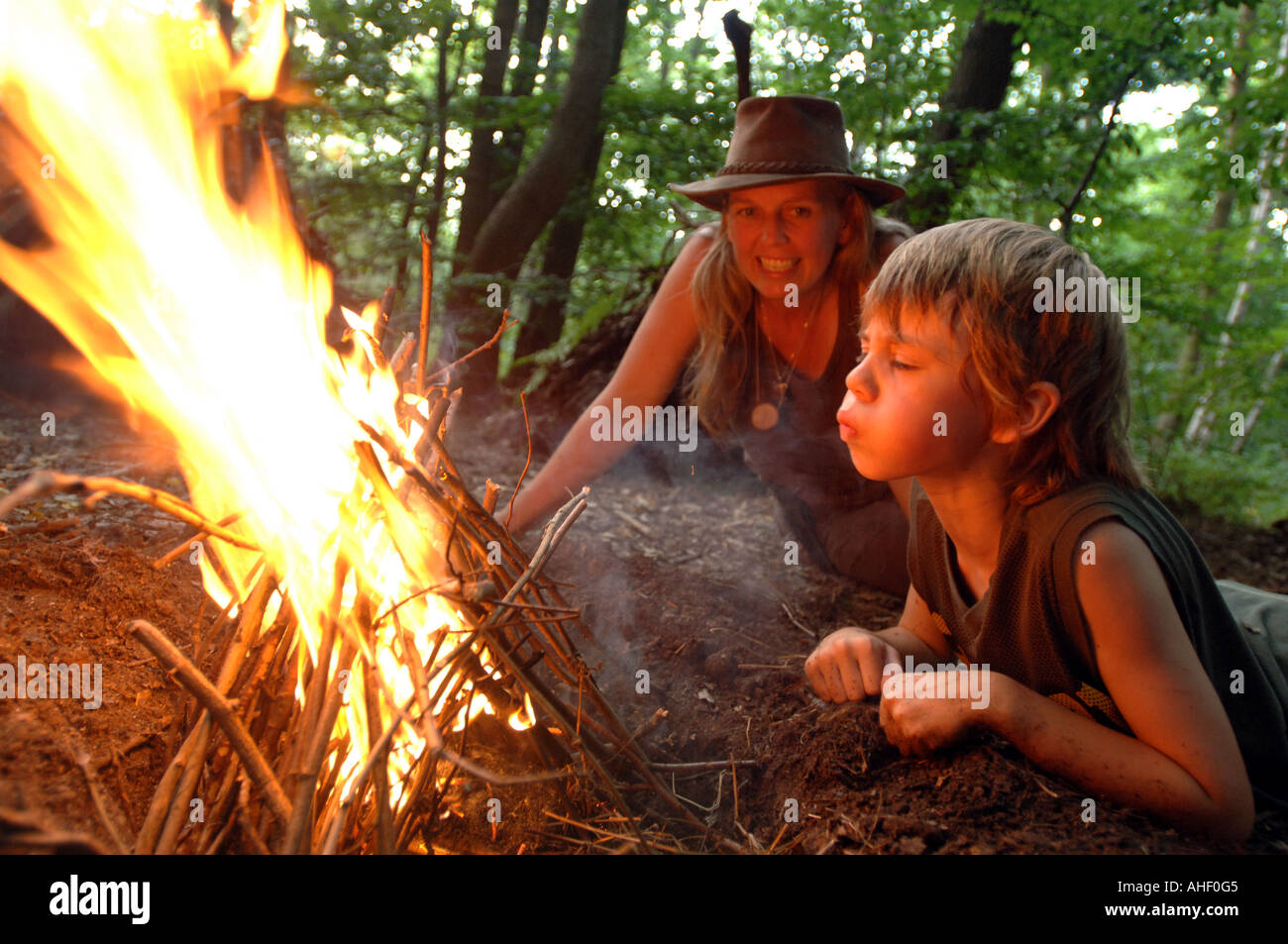 Kleiner Junge bläst einen Lagerfeuer in das Leben in einem Waldgebiet clearing als Frau blickt auf zustimmend Stockfoto